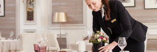 Eine Frau in einem dunkelblauen Kostüm richtet einen Tisch in einem Hotelrestaurant her. Der Tisch ist fein gedeckt mit einer weißen Tischdecke und Blumen.