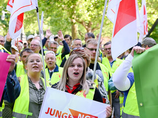 Zwei junge Frauen mit einer Fahne von jungeNGG und neongelben Streikwesten stehen im Vordergrund. Im Hintergrund sieht man eine Menge mit Streikwesten. 