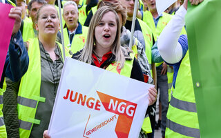 Zwei junge Frauen mit einer Fahne von jungeNGG und neongelben Streikwesten stehen im Vordergrund. Im Hintergrund sieht man eine Menge mit Streikwesten. 